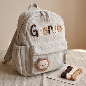 May include: A light grey corduroy backpack featuring the name George embroidered in dark brown, light brown, and cream yarn. A small, circular lion face patch with a brown fringe mane is attached to the front pocket, which has braided zipper pulls.