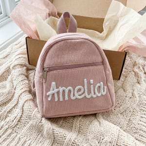 Könnte beinhalten: Ein altrosa Mini-Rucksack aus Kord mit einem Tragegriff und einer Reißverschlusstasche auf der Vorderseite. Der Name Amelia ist in weißer, strukturierter Schreibschrift mittig auf der Vordertasche des kleinen Stoffrucksacks eingestickt.