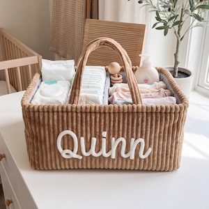 May include: A rectangular brown corduroy storage basket with two handles and the name Quinn embroidered in white script on the front. The interior is divided into sections containing baby wipes, diapers, a wooden rattle, and folded clothing.
