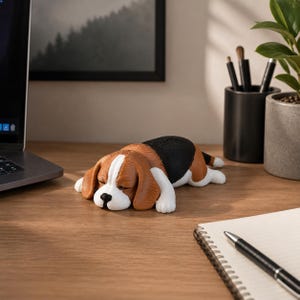 Puede incluir: Una pequeña figura tridimensional de un perro beagle acostado en posición de dormir. El perro presenta un pelaje tricolor con secciones marrones, negras y blancas, un hocico blanco y una nariz negra, elaborado con una textura de capas visible.