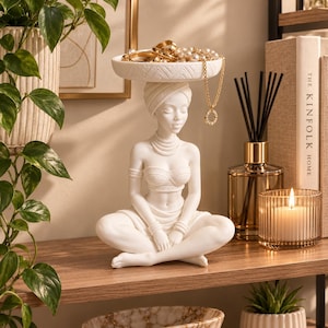 May include: A white resin sculpture of a person sitting in a cross-legged position, wearing a headwrap and jewelry. The figure supports a shallow, textured circular tray on their head, which holds gold-toned rings and a pearl-accented necklace.