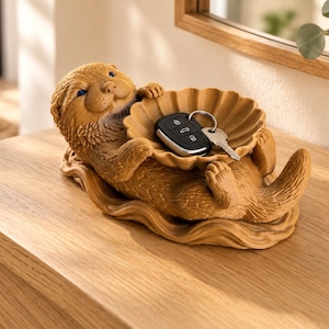 May include: A brown otter-shaped figurine lying on its back, holding a scalloped shell-shaped bowl on its stomach. The bowl contains a black car key fob with a silver metal key attached. The figurine has textured fur, blue eyes, and a wavy base.