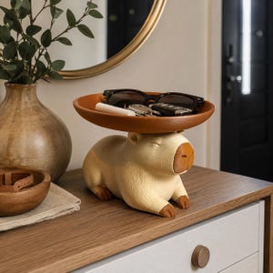 May include: A capybara-shaped figurine featuring a light tan body with a darker brown nose and feet. The figurine supports a round, terracotta-colored tray on its back, which currently holds a pair of black sunglasses, a white tube, and keys.