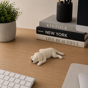 Peut inclure: Une petite figurine couleur crème représentant un chiot endormi couché sur le ventre. Le chiot a une texture lisse, les yeux fermés et un petit nez noir. Il est posé sur une surface en bois devant des livres empilés et un clavier d'ordinateur.