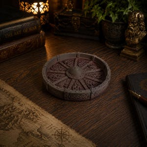 May include: A circular tray featuring a raised rim and a central hub with radiating spokes. The surface is decorated with intricate, embossed Celtic knotwork patterns in a weathered, dark brown finish, resembling a stylized ancient wagon wheel.