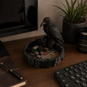 May include: A black raven figurine perched on the edge of a circular, stone-textured tray. The tray features a shield-like emblem on the front and contains several metallic coins and black USB flash drives resting inside the basin.
