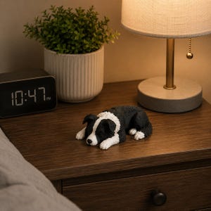 May include: A small, realistic figurine of a Border Collie dog lying down in a resting position. The dog has a black and white coat with a white blaze on its face, a white chest, and white paws. The sculpture is detailed with textured fur.