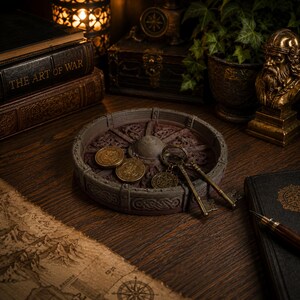 May include: A circular, dark gray stone-like tray featuring intricate Celtic knotwork patterns along the rim and base. Inside the tray are two gold-colored circular coins and two antique-style brass skeleton keys attached to a metal ring.