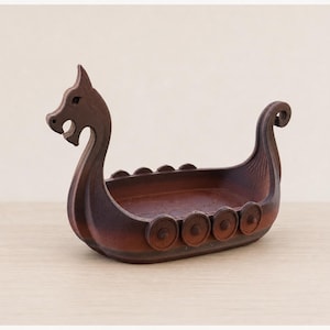 May include: A dark brown wooden bowl shaped like a Viking longship. The vessel features a carved dragon head at the prow, a curled tail at the stern, and circular shield-like details along the sides of the hull, creating a hollow interior.