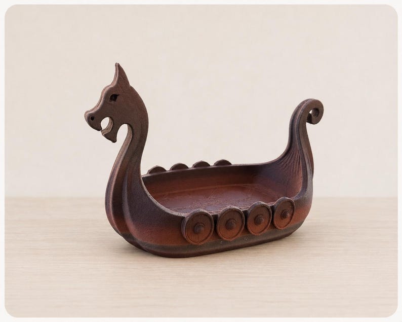 May include: A dark brown wooden bowl shaped like a Viking longship. The vessel features a carved dragon head at the prow, a curled tail at the stern, and circular shield-like details along the sides of the hull, creating a hollow interior.