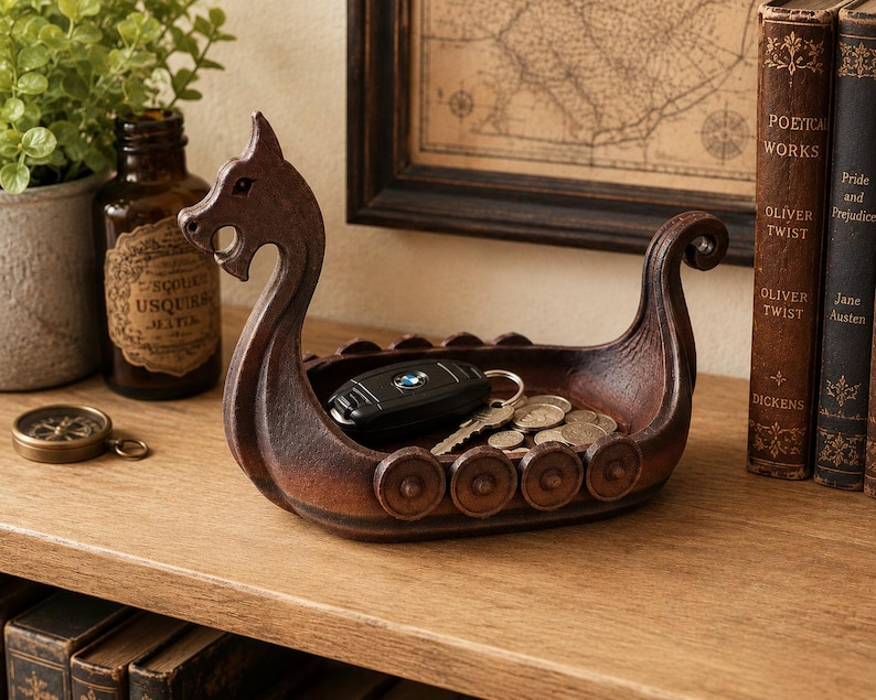 May include: A dark brown, carved-style decorative bowl shaped like a Viking longship. The vessel features a prominent dragon-head prow, a curved stern, and circular shield motifs along the sides. It contains a black car key fob and metal coins.