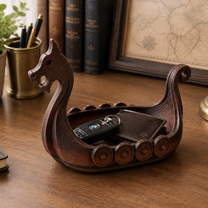 May include: A brown, boat-shaped storage tray designed like a Viking longship with a carved dragon head at the prow and circular shield details along the sides. The interior of the vessel holds a black car key fob and a dark brown leather wallet.