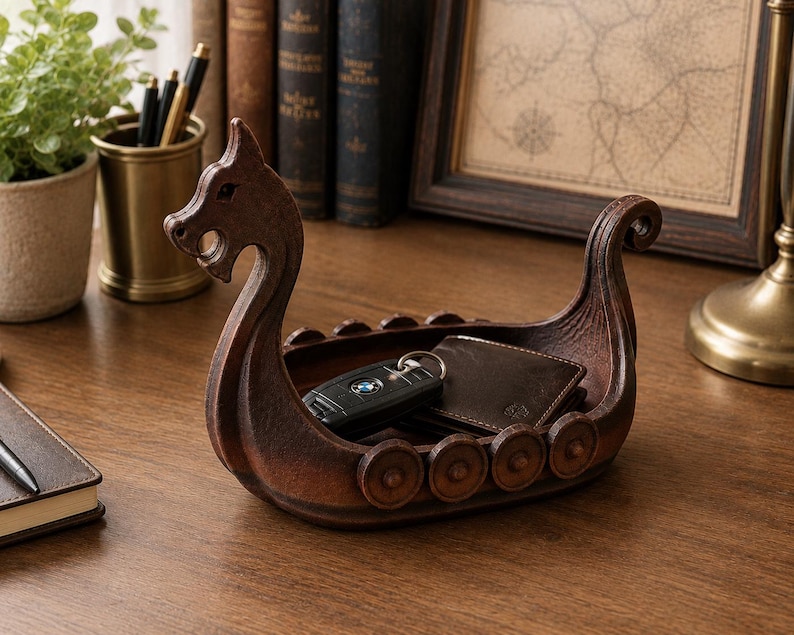 May include: A brown, boat-shaped storage tray designed like a Viking longship with a carved dragon head at the prow and circular shield details along the sides. The interior of the vessel holds a black car key fob and a dark brown leather wallet.
