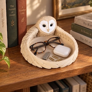 May include: A circular cream-colored trinket tray featuring a sculpted barn owl with outstretched wings forming the rim. The tray holds a pair of tortoiseshell-patterned glasses, a white wireless earbud case, a silver USB drive, and two gold paperclips.