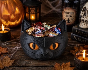 May include: A black cat-shaped bowl with textured fur details and large orange eyes, filled with various wrapped chocolate candies. The bowl features pointed ears and a molded feline face, serving as a container for assorted treats.