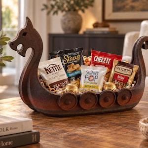 May include: A dark brown, carved wooden bowl shaped like a Viking longship, featuring a dragon-head prow and circular shield details along the sides. The bowl is filled with various snack bags including Kettle Brand, Stacy's, Cheez-It, and SkinnyPop.