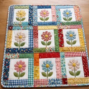 May include: A rectangular fabric quilt featuring a grid of nine white squares, each displaying a single appliquéd flower with a yellow center and green leaves. The squares are framed by various colorful patterned fabric strips in gingham, floral, and solid prints.