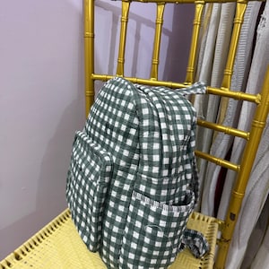 May include: A quilted backpack featuring a dark green and white gingham check pattern. The backpack includes a large main compartment with a zipper closure, a smaller front pocket, and side pockets. The fabric has a visible stitched texture.