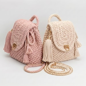 May include: Two crochet backpacks, one in dusty pink and one in cream, featuring a textured knit pattern, a top handle, a fold-over flap with a gold-toned metal clasp, and decorative hanging tassels on the sides with matching chain shoulder straps.