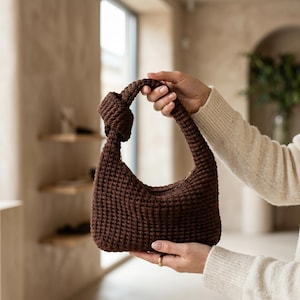 Pode incluir: Uma bolsa de mão castanho-escura feita em crochê, com um corpo estruturado em forma de meia-lua e uma alça única integrada com um detalhe de nó proeminente no topo. A bolsa é feita de fio grosso e texturizado com um padrão de pontos denso.