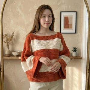 May include: A long-sleeved crochet sweater featuring wide horizontal stripes in burnt orange and cream. The garment has a wide boat neckline and loose, flowing sleeves that taper slightly at the cuffs, crafted with a consistent double crochet stitch.