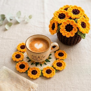 Pode incluir: Uma caneca de cerâmica com café e espuma em forma de coração, sobre um porta-copos redondo de crochê branco decorado com girassóis amarelos e castanhos, ao lado de uma pequena cesta de crochê castanha cheia de girassóis de crochê.