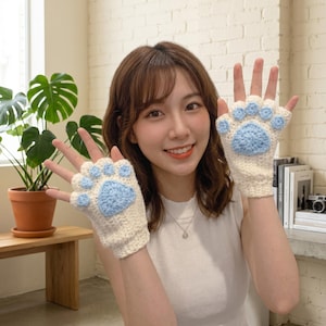 May include: A pair of fingerless crochet gloves in cream white with light blue paw pad accents. The gloves feature a large central triangular pad and four smaller circular pads on the knuckles, mimicking an animal paw design.