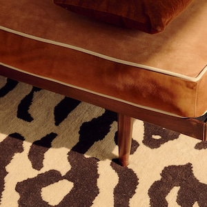 May include: A brown velvet bench with cream-colored piping and a matching brown velvet cushion. The bench features a dark wooden frame and tapered legs, positioned over a cream-colored rug with a large-scale black abstract animal print pattern.
