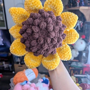 May include: A large, handmade crochet sunflower with a plush texture. The petals are a vibrant yellow, surrounding a textured, dark brown centre. This decorative item is a unique, handcrafted piece.