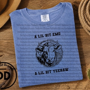 May include: A blue short-sleeved t-shirt featuring a black graphic of a cow split down the middle, with the text A LIL BIT EMO above and A LIL BIT YEEHAW below the graphic. The shirt has a crew neckline and a Comfort Colors brand label at the neck.