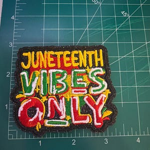 May include: Embroidered patch with the words "JUNETEENTH VIBES ONLY" in yellow, green, and red. The patch has a black border with glitter and is on a green cutting mat.