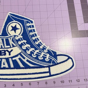 May include: A blue and white embroidered patch shaped like a high-top sneaker. The design features white laces, a circular star emblem, and the text WALK BY FAITH in bold blue letters. The patch is positioned on a purple cutting mat with inch markings.