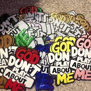 May include: A collection of embroidered patches featuring the text "GOD DON'T PLAY ABOUT ME" in various colors including red, yellow, pink, blue, green, gold, and white. Each patch has a textured surface with bold, block-style lettering.
