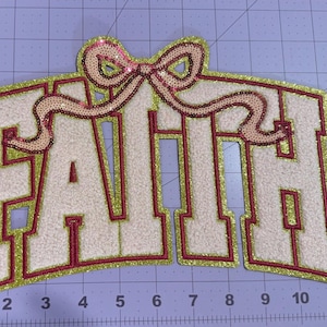 May include: A large iron-on patch featuring the word FAITH in white chenille letters with a red border and a gold glitter outline. A pink sequined bow sits atop the letters. The patch is positioned on a cutting mat with inch markings.