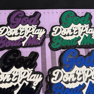 May include: Four embroidered patches featuring the text God Don't Play Bout Me! in a script font. The patches come in purple, green, black, and blue, each with white lettering for the middle line and a glittery finish on the colored text.