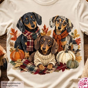 May include: A cream-colored t-shirt featuring a graphic of three dachshunds wearing scarves and a sweater, surrounded by autumn leaves and pumpkins. The dogs are positioned centrally, with one wearing a plaid scarf, one a knit scarf, and one a sweater.