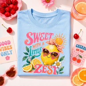 May include: Light blue t-shirt with a lemon graphic wearing sunglasses and a bow, the text reads "Sweet with a little zest". The image includes cherries, orange slices, and the text "Good Vibes Only" and "Cherry on Top".