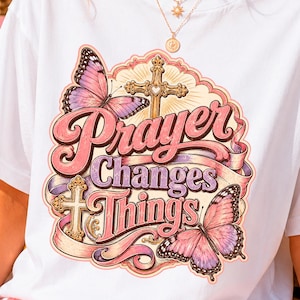 Peut inclure: Un t-shirt blanc avec une impression graphique portant le texte "Prayer Changes Things" en lettres roses et violettes. Le motif comprend deux croix dorées ornées, deux grands papillons violets et roses, et des rubans décoratifs.