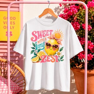 May include: White t-shirt with a colorful graphic of a lemon wearing sunglasses and a bow, with the words "Sweet with a Little Zest". The design includes a sun, flowers, and lemon slices. The shirt is hanging on a wooden hanger.