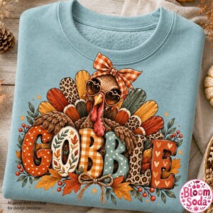 May include: A light blue crewneck sweatshirt featuring a graphic of a turkey wearing a plaid bow and heart-shaped glasses. The turkey is surrounded by colorful feathers and autumn leaves. The word GOBBLE is printed in large, patterned letters.
