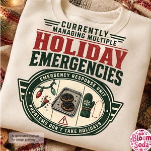 May include: A cream-colored sweatshirt featuring the text "Currently Managing Multiple Holiday Emergencies" in red and dark green. The graphic includes a central emblem with a baking sheet of cookies, a green oven mitt, and broken light bulbs.