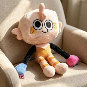 May include: A plush toy with a square-shaped peach head, large black and white eyes, and a small smile. It features black arms, one blue hand with pink claws, one pink hand with blue claws, and peach legs with orange stripes on a yellow torso.
