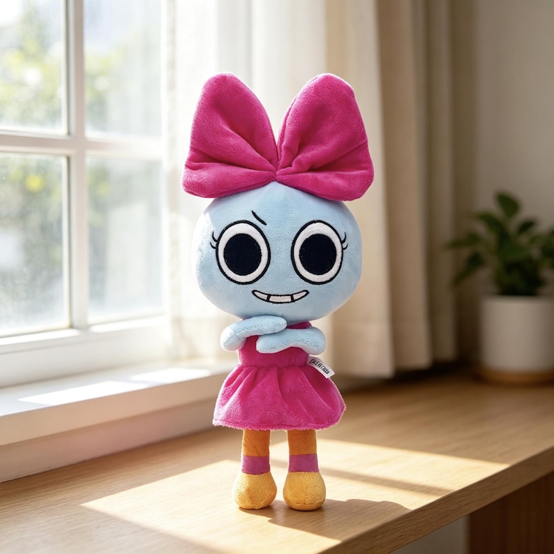 May include: A plush doll featuring a light blue round head with large black eyes and a small grin. It wears a large bright pink bow on top of its head, a matching pink dress, and orange legs with pink bands above yellow feet. A small white tag is visible.