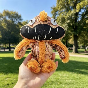 May include: A plush toy featuring a textured orange and brown patterned body with long orange faux fur accents. It has a large black mouth area with white teeth, two small white circular eyes with dark pupils, and a small tuft of orange fur on top.