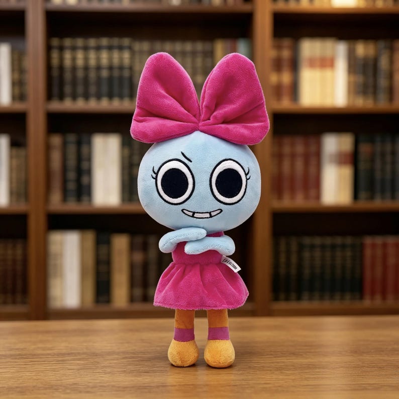 May include: A plush toy featuring a light blue round head with large black eyes, a small grin, and a prominent bright pink bow on top. The figure wears a matching pink dress, orange legs, and pink socks, with arms crossed over its chest.