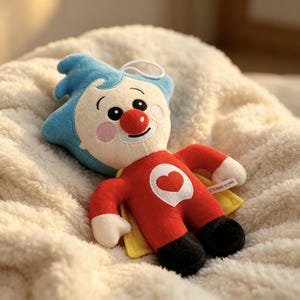 May include: A plush toy featuring a character with blue hair, a red nose, and pink cheeks. The figure wears a red outfit with a white heart emblem on the chest, black feet, and a yellow cape. A small white tag is attached to the side of the body.