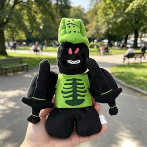 May include: A plush toy featuring a black character with a green helmet, a green torso with a dark green ribcage graphic, and two black swords. The character has pink eyes and a white mouth with sharp teeth, and is constructed from soft fabric.