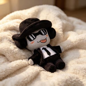 May include: A plush doll featuring a light gray face with embroidered black sunglasses, a small orange tongue, and pink cheeks. The doll wears a black hat, a black suit jacket, a white shirt, and a black tie, with matching black pants and shoes.