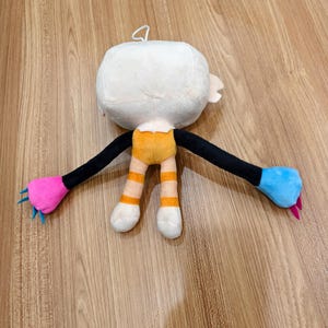 May include: A plush toy featuring a large white head, a yellow torso, and striped yellow and white legs. The long black arms end in one bright pink hand with blue claws and one bright blue hand with pink claws. A small white loop is on the head.