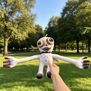 May include: A white plush toy with a large head, wide brown eyes, and a wide mouth filled with sharp teeth. The creature has long, thin arms and legs with dark purple tips on the hands and feet, and a small dark brown collar around its neck.